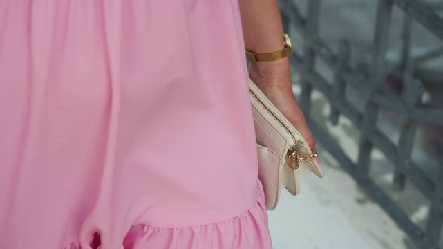 white woman pink dress holding clutch, standing by iron railing, rose gold clutch and gold bracelet, flowing ruffle hem, soft daylight, tactile fabric texture, editorial romantic mood, feminine