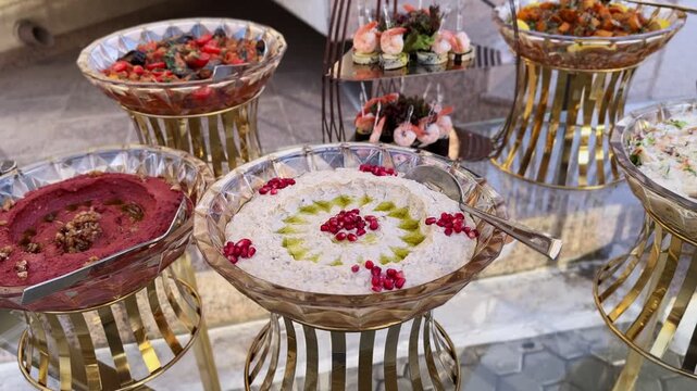 Close-up of elegant Arabic mezze buffet: baba ghanoush with olive oil and pomegranate, muhammara, mixed salads and shrimp canap&eacute;s on gold stands&mdash;perfect for catering and events.