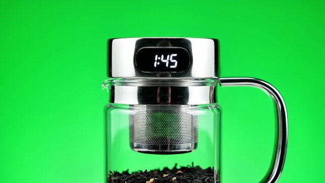 A close-up of a glass tea infuser with a digital timer on a bright green background