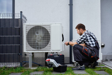 Technician performing maintenance on outdoor heat pump unit near residential building exterior. Man installing air source heat pump, using tools. Concept of HVAC service and heating system repair