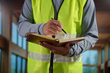 A professional construction engineer or site supervisor stands confidently in a modern wooden interior, wearing a high-visibility safety vest and holding rolled architectural blueprints