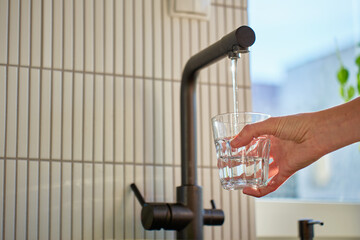 Hand filling glass with drinking water from kitchen faucet in modern home interior. Woman pouring...