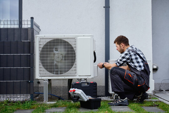 Technician performing maintenance on outdoor heat pump unit near residential building exterior. Man installing air source heat pump, using tools. Concept of HVAC service and heating system repair