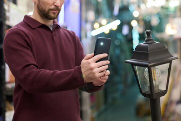 Caucasian young adult man using smartphone to photograph outdoor lamp post in hardware store, standing with focused expression, holding device with both hands, shelves visible in background