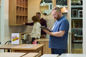 Caucasian man using digital tablet in furniture store while two Caucasian adults browsing kitchen cabinets in background during super sale event