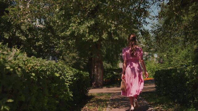 caucasian woman walking down sunlit path through green hedges and oak trees, wearing flowing pink dress, backshot evokes romantic solitude and serene summer mood, frames suggest model, florist,