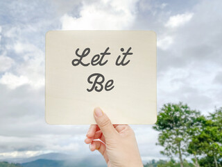 Hand holding a card with the words Let It Be against a cloudy sky and nature background, representing peace, acceptance and mindfulness.