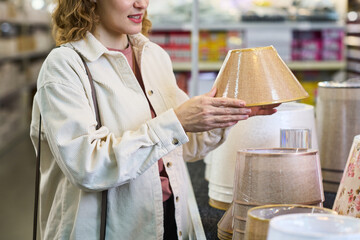 Caucasian young adult woman examining lampshade while shopping in store, holding item with both hands and smiling, standing near display with various lighting products visible