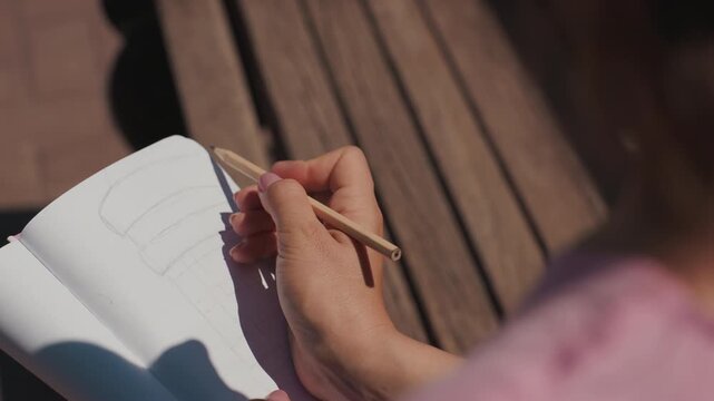 Closeup hands sketching notebook on bench, woman uses pencil to draw simple outline warm sunlight, wooden slats, soft shadow, focus on tactile movement, intimate reflective mood, creative study