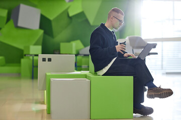 Caucasian young adult man sitting on modern green cube furniture using laptop and holding reusable coffee cup in contemporary workspace with geometric wall decor