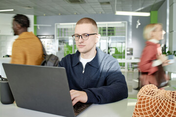 Caucasian young adult man wearing glasses working on laptop in modern office space, focused on screen while diverse people walking in background, hands typing on keyboard