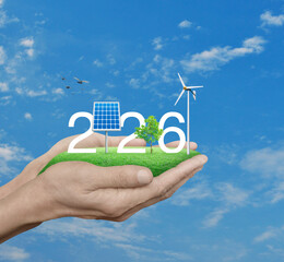 2026 white text with solar cell, wind turbine and growing tree on green grass field in hands over blue sky, white clouds and birds, Happy new year 2026 ecology saving power and energy concept