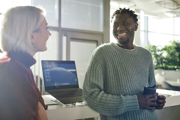 Young adult Caucasian woman and young adult Black man standing in modern office, smiling and talking near laptop, man holding reusable coffee cup, natural light coming through windows