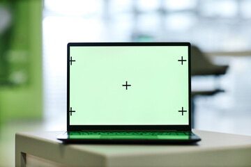 Laptop computer with blank green screen displaying alignment marks standing on table in modern office environment, screen ready for chroma key compositing or digital content insertion