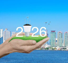 2026 white text with wind turbines on soil and light bulb on green grass in hands over city tower, skyscraper and river with birds, Happy new year 2026 green ecology and saving energy concept