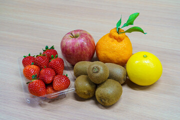Assortment of fresh fruits on a wooden table. Strawberries in a plastic pack, red apple, kiwi fruits, Shiranui citrus and yellow orange. Healthy dessert ingredients.