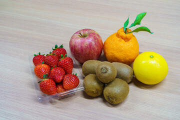 Assortment of fresh fruits on a wooden table. Strawberries in a plastic pack, red apple, kiwi fruits, Shiranui citrus and yellow orange. Healthy dessert ingredients.