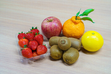 Assortment of fresh fruits on a wooden table. Strawberries in a plastic pack, red apple, kiwi fruits, Shiranui citrus and yellow orange. Healthy dessert ingredients.