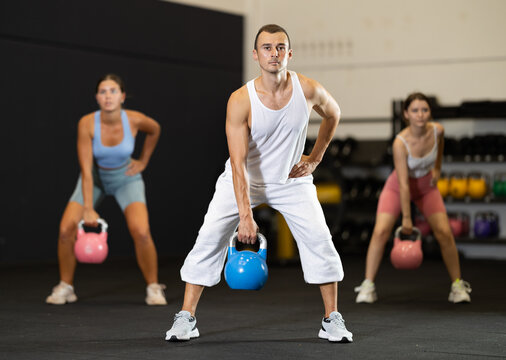 Sportive strong male in activewear swinging kettlebell during group exercise class in crossfit gym indoors - Powered by Adobe
