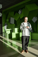 Young adult Caucasian man standing indoors using smartphone, looking down at device with neutral...