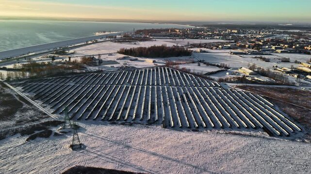 Drone aerial of a snow-dusted solar farm near the coastline, with long photovoltaic panel rows stretching across frozen fields beside a winter town.