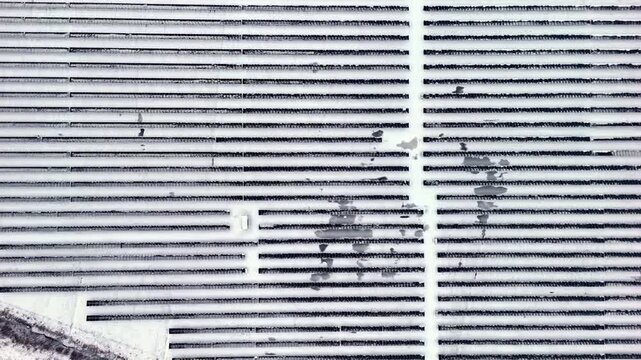 Top-down drone view of a snow-covered solar panel array in neat parallel rows, creating an abstract winter geometry of renewable power infrastructure.