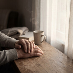 Close-up of older hands resting on rustic wooden table near steaming coffee mug and book next to sunlit window