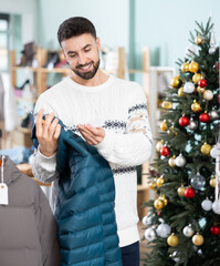 Young man buyer choosing warm down jacket in clothing store decorated for Christmas