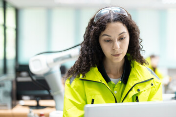 Female engineer in high-visibility jacket working on a laptop with robotic arm in the background, representing smart factory operations, digital automation and modern industrial technology.