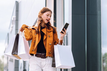A young woman stands outside a contemporary building,checking her phone while holding shopping bags. She appears excited as she arranges for her taxi.Purchases, black friday, discounts, sale concept.