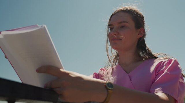 Young white woman reading script outdoors under bright sky, sun backlighting wet hair, pink blouse, holding stack of pages, focused expression, wristwatch visible on arm, quiet rehearsing moment