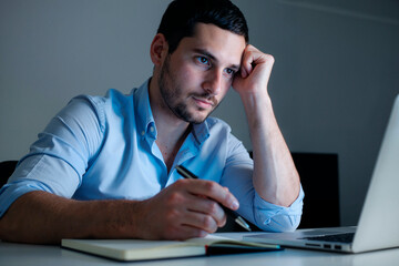 Caucasian young adult man sitting at desk holding pen, looking at laptop screen with thoughtful expression, resting head on hand, notebook open in front, appearing focused on work