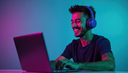 Young man with headphones laughs typing on laptop. Wears headphones, works plays on computer with neon lighting. Tech savvy guy enjoys online activity entertainment. Teenager gamer smiles with