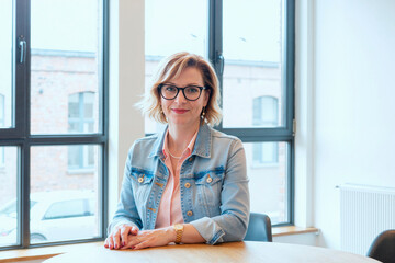 Portrait of Caucasian middle aged woman with short blonde hair and glasses sitting at table smiling confidently hands folded in modern office setting large windows in background