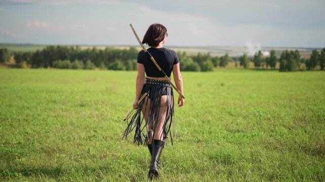 caucasian female warrior walking across grassland nomadic scout moving toward distant horizon with spear strapped to back leather skirt tassels swaying heavy boots pressing into sunlit meadow
