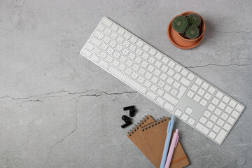 Horizontal top view of modern desk with keyboard, notebooks, pastel pens and wireless earbuds on gray stone background with copy space. Concept of advertising, marketing and social media