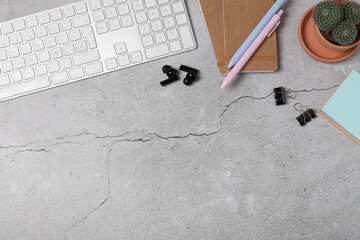 Horizontal flat lay of modern workspace with keyboard, notebook, pastel pens, wireless earbuds, cactus and sticky notes on gray concrete background with copy space. Concept of marketing