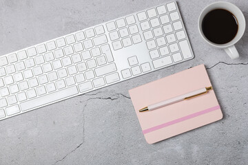 Horizontal top view of modern desk with white keyboard, pink notebook, pen and cup of coffee on gray stone background. Copy space. Concept of productivity, office lifestyle and remote work