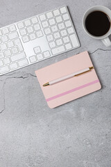 Vertical top view of minimalist workspace with white keyboard, pink notebook, pen and coffee cup on gray stone surface. Copy space. Concept of modern workspace and creative planning