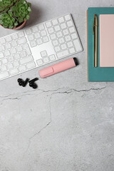 Top view vertical composition of minimalist workspace with white keyboard, pink notebook, pen, wireless earbuds and succulent on gray stone surface. Copy space. Concept of digital lifestyle