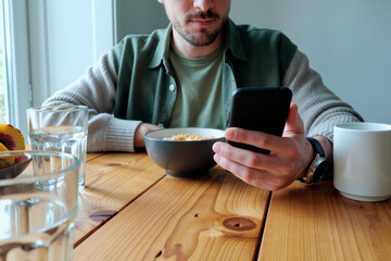 Young adult Caucasian man sitting at wooden table holding smartphone in right hand while eating breakfast cereal from bowl, focusing on device screen, partial face visible