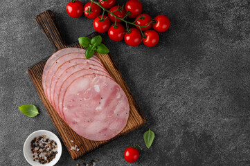 Delicious ham cut into round slices on a cutting board with spices, cherry tomatoes and basil leaves on a dark stone background. Top view.