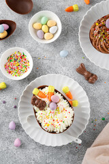 Easter dessert. Chocolate eggs with cheesecake filling decorated with chocolate candies and sugar sprinkles on a white plate on gray concrete background. Copy space.