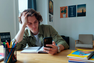 Caucasian teenage boy sitting at desk holding smartphone in one hand while looking at screen with thoughtful expression, open notebook and colorful folders on table