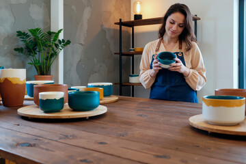 Caucasian young adult woman examining ceramic bowl while standing behind wooden table with various handmade pottery pieces displayed on round trays in modern studio