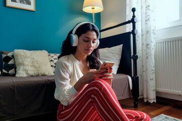 Caucasian young adult woman sitting on floor using smartphone while wearing wireless headphones, relaxing in modern bedroom, focused on screen, natural light from window