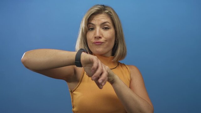 Woman wearing orange sleeveless top pointing finger to watch on bare wrist in blue studio; impatience time pressure urgency.