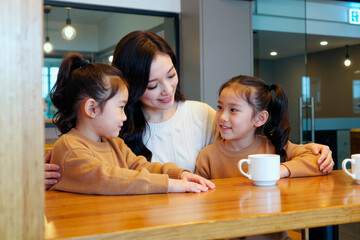 Asian young adult woman sitting at table embracing two Asian girls, both children smiling and interacting with each other, coffee mugs placed in front of them on wooden surface