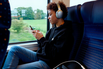 Young adult Black woman sitting on train seat listening to music with headphones while using smartphone, looking at device screen, traveling during daytime with green landscape outside window
