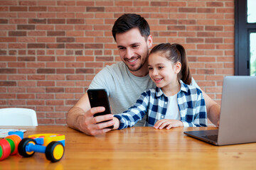 Caucasian young adult man sitting with Caucasian child girl smiling and looking at smartphone together at table with laptop, both engaging with device in modern home setting